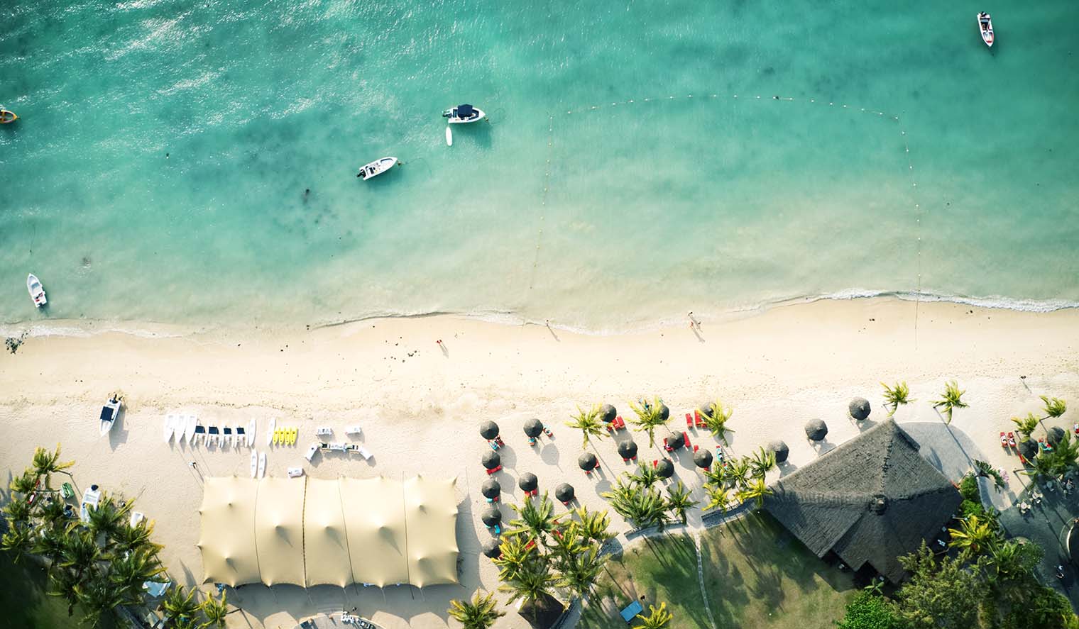 Luftaufnahme eines Traumstrandes auf Mauritius mit türkisfarbenem Wasser, Booten, Palmen, Sonnenschirmen und entspannter Ferienatmosphäre – ideal für Mauritius Badeferien